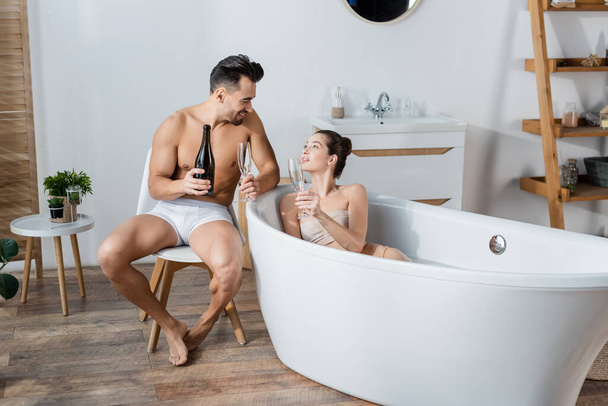 young woman in bathtub and sexy man in underpants holding champagne glasses and smiling at each other - Фото, зображення