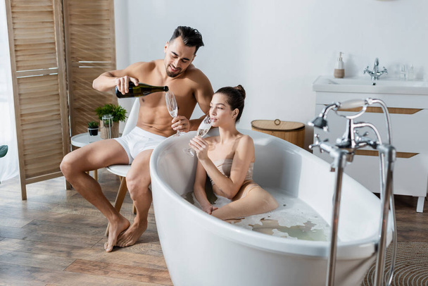 uśmiechnięty mężczyzna w bieliźnie nalewający szampana obok dziewczyny kąpiący się w przestronnej łazience - Zdjęcie, obraz