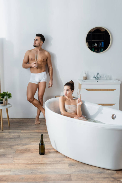 sexy man in underpants standing with champagne glass near girlfriend relaxing in bathtub - Фото, изображение