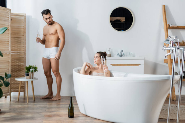 smiling woman relaxing in bathtub near boyfriend in underpants standing with champagne glass - 写真・画像