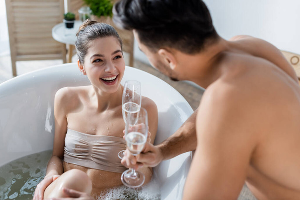 veselá žena se koupe a dívá se na rozmazaný přítel v blízkosti sklenice šampaňského - Fotografie, Obrázek