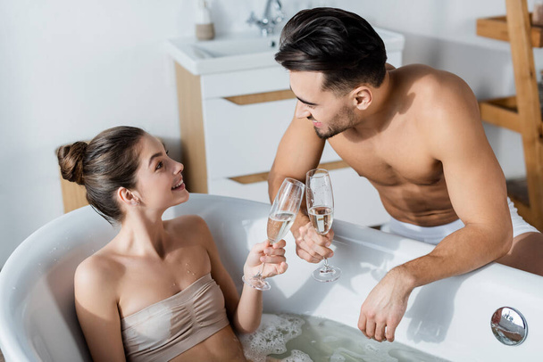 joyful woman in bathtub clinking champagne glasses with sexy muscular boyfriend - Photo, Image