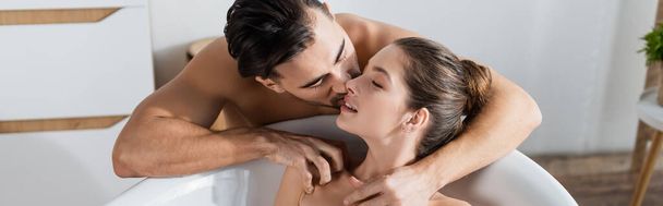 shirtloze man zoenen lachende verleidelijke vriendin ontspannen in bad thuis, banner - Foto, afbeelding