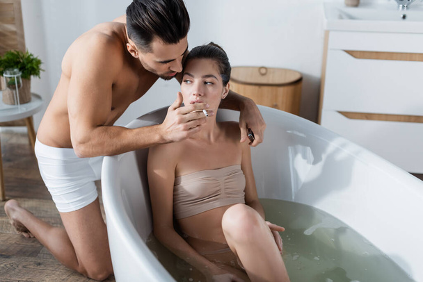 sexy man in onderbroek houden sigaret in de buurt sensuele vrouw nemen bad in huis - Foto, afbeelding