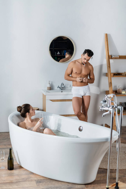sexy man in underpants lighting cigarette near girlfriend relaxing in bathtub with glass of champagne - Фото, зображення