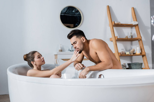 соблазнительная женщина, держащая сигарету рядом с сексуальным парнем без рубашки, расслабляясь в ванной с бокалом шампанского - Фото, изображение