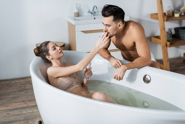 sexy woman relaxing in bathtub with glass of champagne and holding cigarette near boyfriend - Фото, изображение