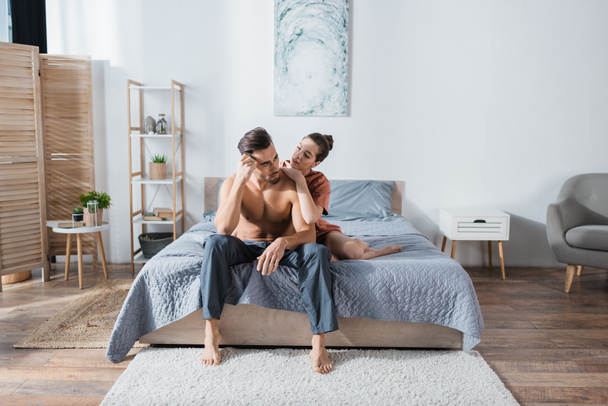 young woman embracing shirtless boyfriend sitting in pajama pants in spacious bedroom - Photo, Image