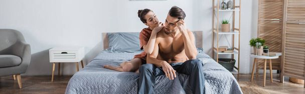 sexy muž v pyžamu kalhoty blízko přítelkyně objímající ho na posteli doma, prapor - Fotografie, Obrázek