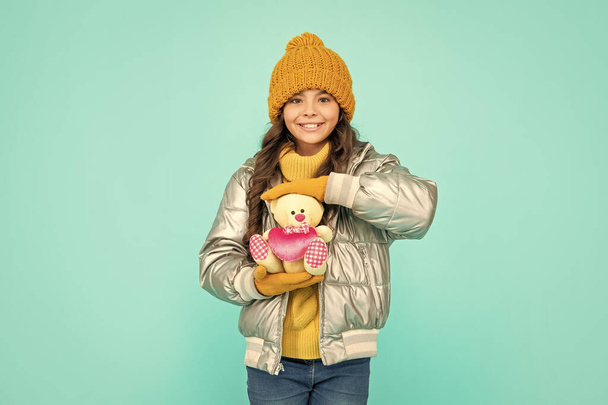 winter fashion. love toy for valentines day. happy kid in puffer jacket and hat. childhood. teen girl showing toy bear. child wearing warm clothes on blue background. express positive emotion. - Zdjęcie, obraz