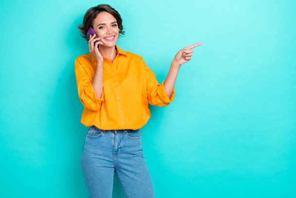 Portrait photo of young cute nice girl wear orange shirt talking phone positive smile finger directing empty space isolated on cyan color background. - Φωτογραφία, εικόνα
