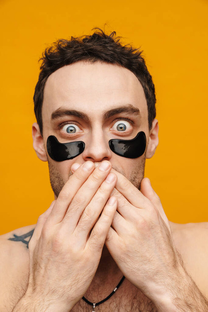 Close up portrait of a shocked brunette mid aged shirtless man wearing cosmetic eye patches standing over yellow wall, cover mouth - Фото, изображение