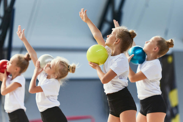 Teamwork at sports. Group of kids, little girls, beginner gymnastics athletes in sports uniform at training at sports gym, indoors. Concept of beauty, sport, achievements, studying, goals, skills - Фото, изображение