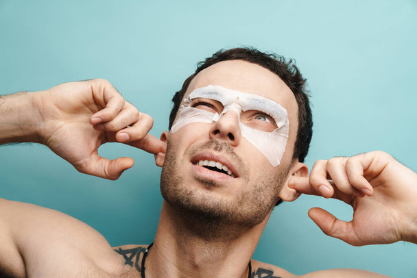 Close up of a mid aged white shirtless man with cosmetic face mask standing over blue background blocking ears - Φωτογραφία, εικόνα