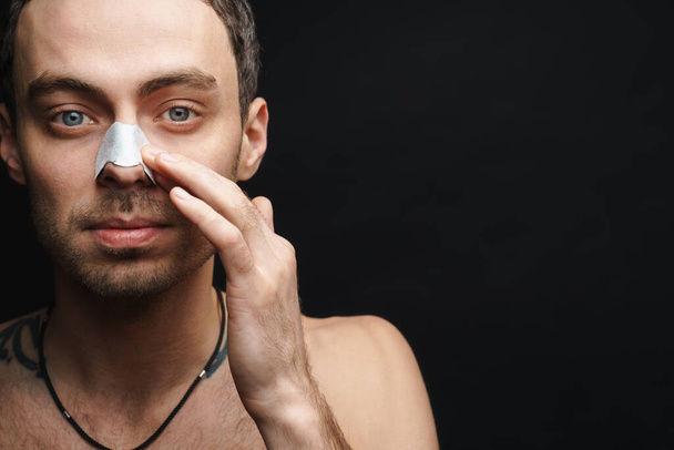 Mid aged white shirtless man using cleansing strip for nose over black wall background - Fotografie, Obrázek