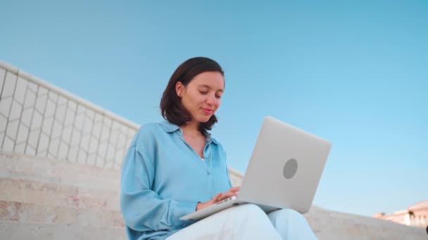 Üzletasszony használja laptop kültéri ülő lépcső kék ég háttér. Fehér nő üzletember 30 éve gépel laptop billentyűzetet. Távoli munka szabadúszó koncepció. Szabadúszó munka az új normális - Felvétel, videó