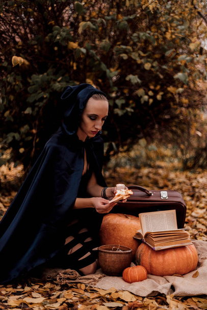 Die Hexe sitzt auf dem Boden auf herbstgelben Blättern. Vor ihr liegen Kürbisse, ein Buch, eine Kerze und ein Koffer. Sie zaubert. - Foto, Bild