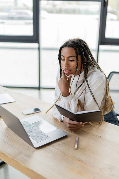 African american businesswoman holding notebook near laptop and blurred smartphone in office  - Foto, Imagem