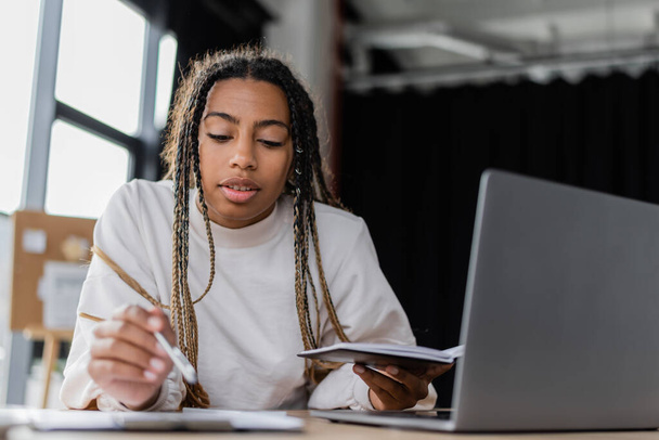 African american businesswoman holding notebook and blurred pen near devices in office - Фото, зображення