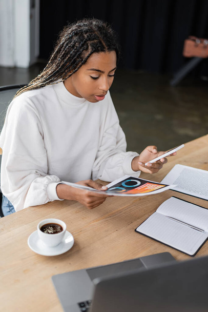 African american businesswoman using smartphone and holding paper with charts near coffee in office  - 写真・画像