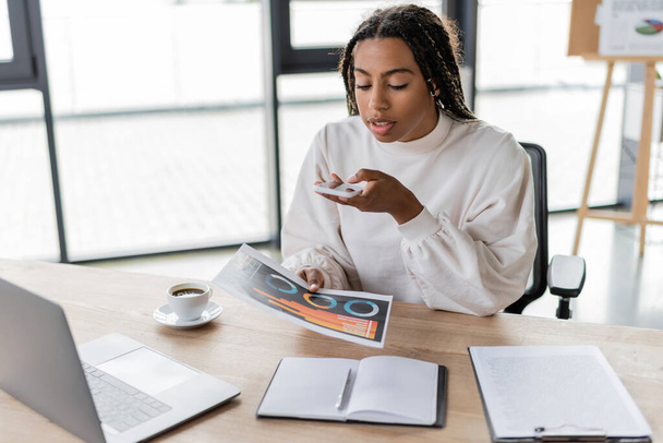 African american businesswoman taking photo of charts near laptop and coffee in office  - Foto, Bild