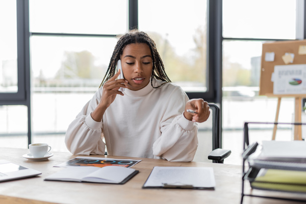 African american businesswoman talking on cellphone near charts and coffee in office  - Photo, Image