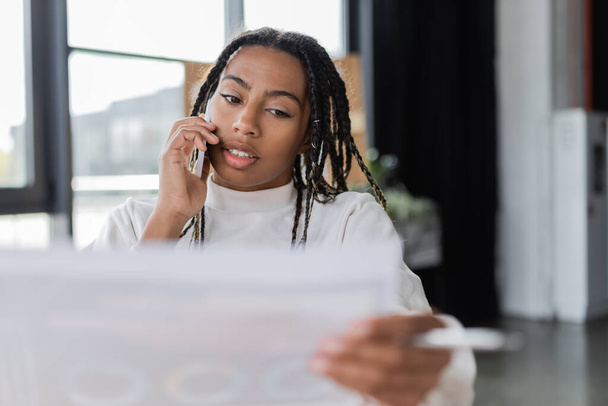African american businesswoman holding blurred paper and talking on smartphone in office  - Foto, imagen