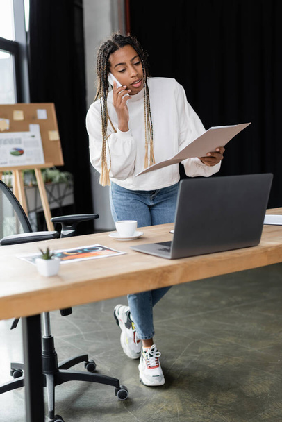 African american businesswoman talking on smartphone and holding paper folder near laptop and coffee in office  - Valokuva, kuva