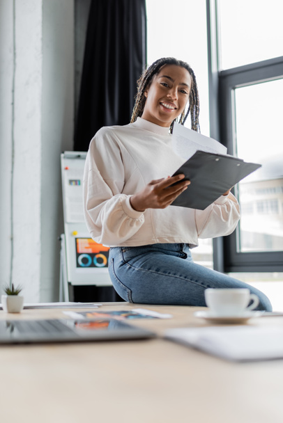 Positive african american businesswoman in casual clothes holding clipboard and looking at camera in office  - Фото, зображення