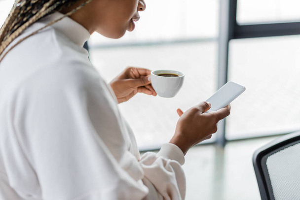 Gehakte weergave van Afrikaanse Amerikaanse zakenvrouw met kopje koffie en het gebruik van smartphone in het kantoor  - Foto, afbeelding