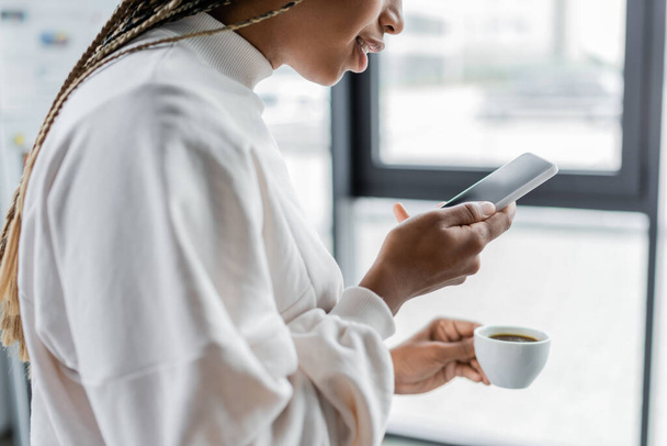Gekropte weergave van Afrikaanse Amerikaanse zakenvrouw met smartphone en koffiekop in het kantoor  - Foto, afbeelding