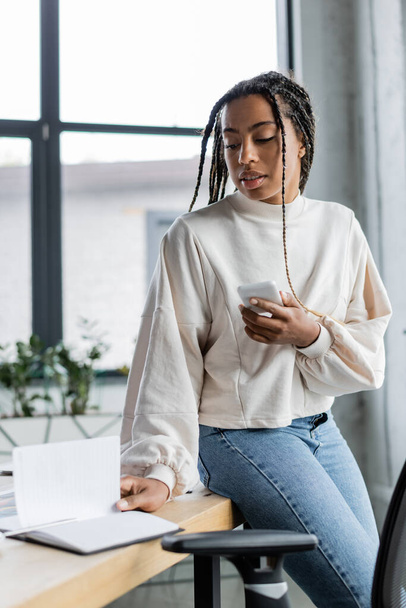 African american businesswoman holding smartphone and looking at notebook in office  - Photo, Image