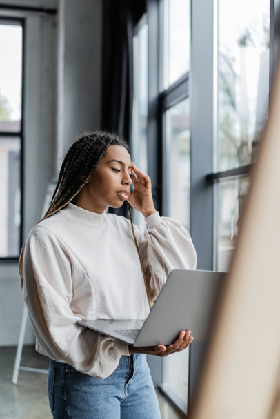 Focused african american businesswoman in sweatshirt holding laptop in office  - 写真・画像