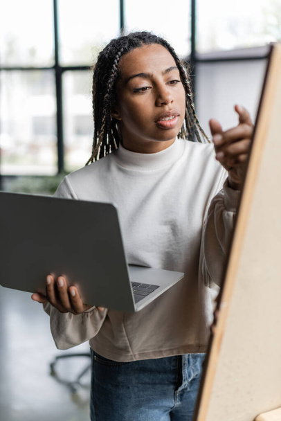 African american businesswoman holding laptop near blurred board in office  - 写真・画像