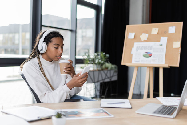 Mujer de negocios afroamericana en auriculares con café para llevar y el uso de teléfonos inteligentes en la oficina  - Foto, Imagen