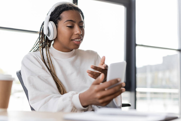 Smiling african american businesswoman in headphones having video chat on smartphone near coffee in office - Zdjęcie, obraz
