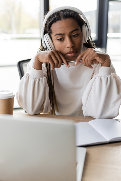 African american businesswoman in headphones holding pen near notebook and blurred laptop in office  - Φωτογραφία, εικόνα