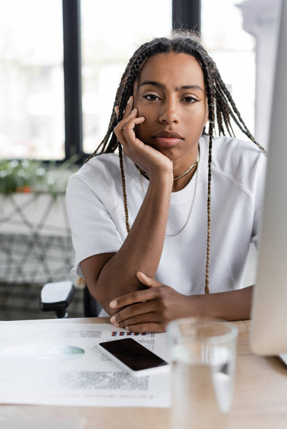 African american businesswoman in t-shirt looking at camera near devices and blurred glass of water  - Fotó, kép