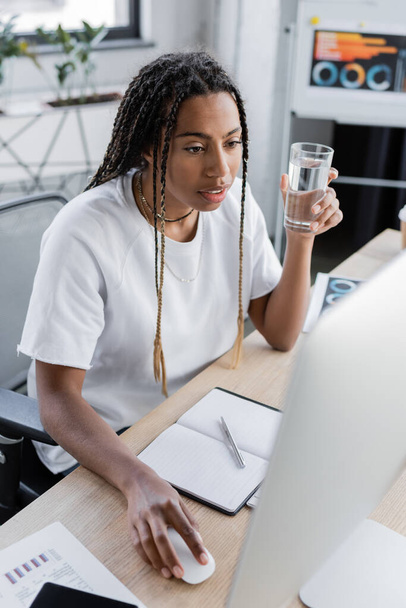 African american businesswoman holding glass of water and using computer in modern office  - Foto, imagen