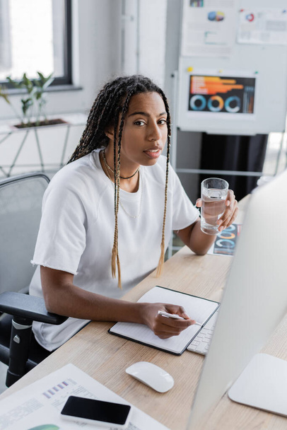 Ofiste bilgisayar ve defterin yanında bir bardak su tutan Afrikalı Amerikalı iş kadını.  - Fotoğraf, Görsel