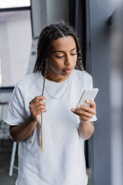 Donna d'affari afroamericana in t-shirt con smartphone in ufficio sfocato  - Foto, immagini