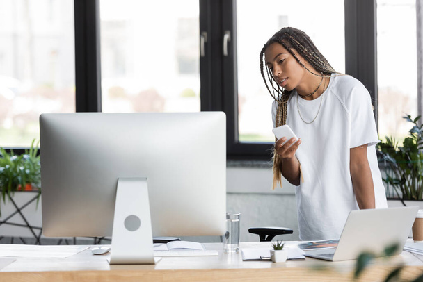 African american businesswoman in t-shirt holding smartphone near computers and papers in office - Zdjęcie, obraz