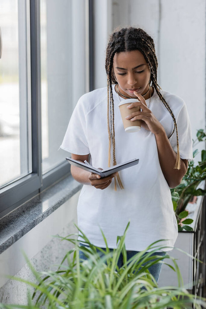 African american businesswoman holding coffee to go and notebook near plants in office  - 写真・画像