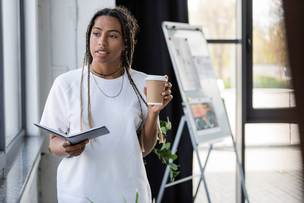 African american businesswoman holding notebook and takeaway drink in office  - Foto, Bild