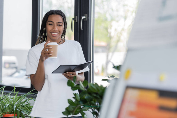 Positive african american businesswoman in t-shirt holding takeaway drink and notebook in office  - Fotografie, Obrázek