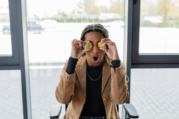 KYIV, UKRAINE - APRIL 27, 2022: Shocked african american businesswoman holding bitcoins near eyes in office  - Photo, Image