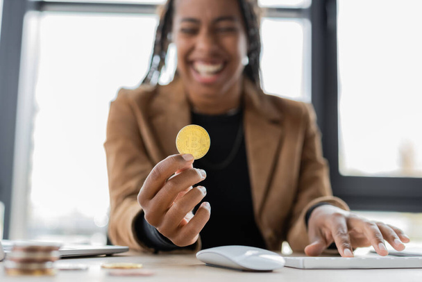 KYIV, UKRAINE - APRIL 27, 2022: Blurred african american businesswoman holding bitcoin near computer keyboard in office  - Photo, Image