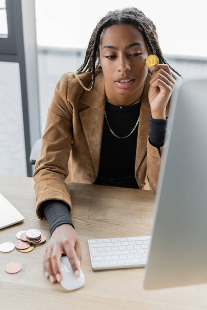 KYIV, UKRAINE - APRIL 27, 2022: African american businesswoman holding bitcoin and using computer in office  - Fotografie, Obrázek