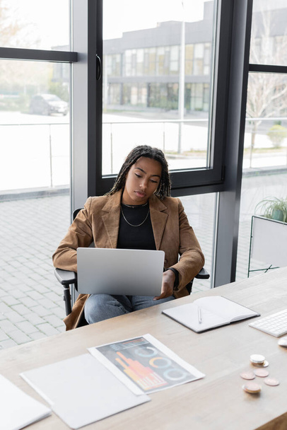 KYIV, UKRAINE - APRIL 27, 2022: African american businesswoman in jacket using laptop near papers and notebook in office  - 写真・画像