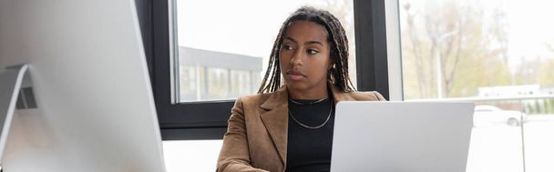 KYIV, UKRAINE - APRIL 27, 2022: African american businesswoman looking at computer monitor near laptop in office, banner  - Foto, Imagem